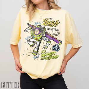 Pode incluir: Camiseta amarelo claro com um desenho de Buzz Lightyear de Toy Story. O design inclui o texto "Buzz Lightyear" e "Ao infinito e al&eacute;m" em uma fonte divertida, com uma ilustra&ccedil;&atilde;o do personagem em seu traje espacial.