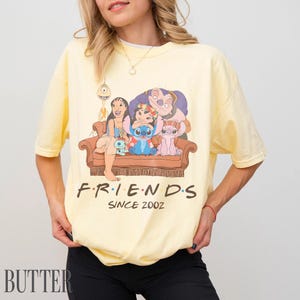 May include: Pale yellow t-shirt with a graphic of Lilo & Stitch characters on a brown sofa. The text "FRIENDS SINCE 2002" is printed below. The shirt has a crew neck and short sleeves. The overall design is reminiscent of the TV show Friends.