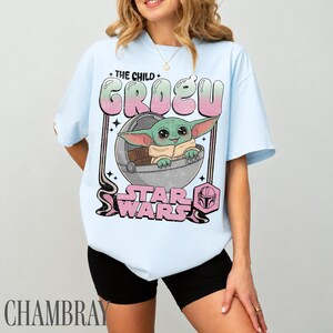 May include: A light blue t-shirt featuring a graphic print of the character Grogu in a floating pod. The text reads THE CHILD GROGU and STAR WARS in a pink and green gradient. A small pink helmet icon is included in a hexagon shape below the text.
