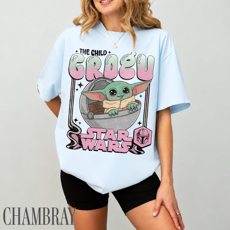 May include: A light blue t-shirt featuring a graphic print of the character Grogu in a floating pod. The text reads THE CHILD GROGU and STAR WARS in a pink and green gradient. A small pink helmet icon is included in a hexagon shape below the text.