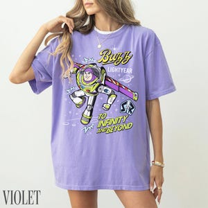 Pode incluir: Camiseta violeta com um desenho de Buzz Lightyear de Toy Story. O design inclui o texto "Buzz Lightyear" e "To Infinity and Beyond". A camiseta tem mangas curtas e um corte folgado.