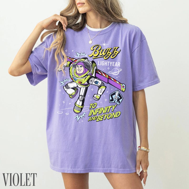 Pode incluir: Camiseta violeta com um desenho de Buzz Lightyear de Toy Story. O design inclui o texto "Buzz Lightyear" e "To Infinity and Beyond". A camiseta tem mangas curtas e um corte folgado.