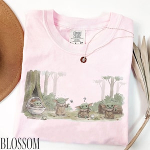 Pu&ograve; includere: T-shirt Comfort Colors rosa chiaro con una grafica di Grogu in una scena di foresta. L'illustrazione mostra Grogu in varie pose, con la parola "Grogu" sulla maglietta. &Egrave; visibile anche una delicata collana con un ciondolo.