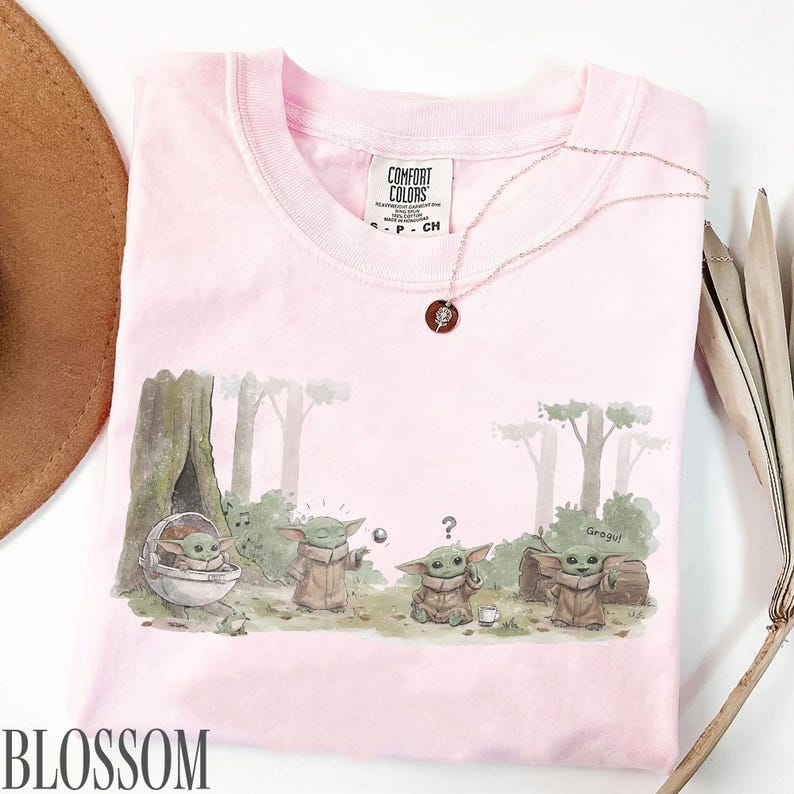 Pu&ograve; includere: T-shirt Comfort Colors rosa chiaro con una grafica di Grogu in una scena di foresta. L'illustrazione mostra Grogu in varie pose, con la parola "Grogu" sulla maglietta. &Egrave; visibile anche una delicata collana con un ciondolo.
