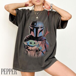 Puede incluir: Una camiseta de manga corta gris carb&oacute;n con un estampado gr&aacute;fico de un guerrero con casco en tonos azules y rojos, junto a una peque&ntilde;a criatura verde con t&uacute;nica beige. El dise&ntilde;o tiene una est&eacute;tica desgastada de estilo vintage.