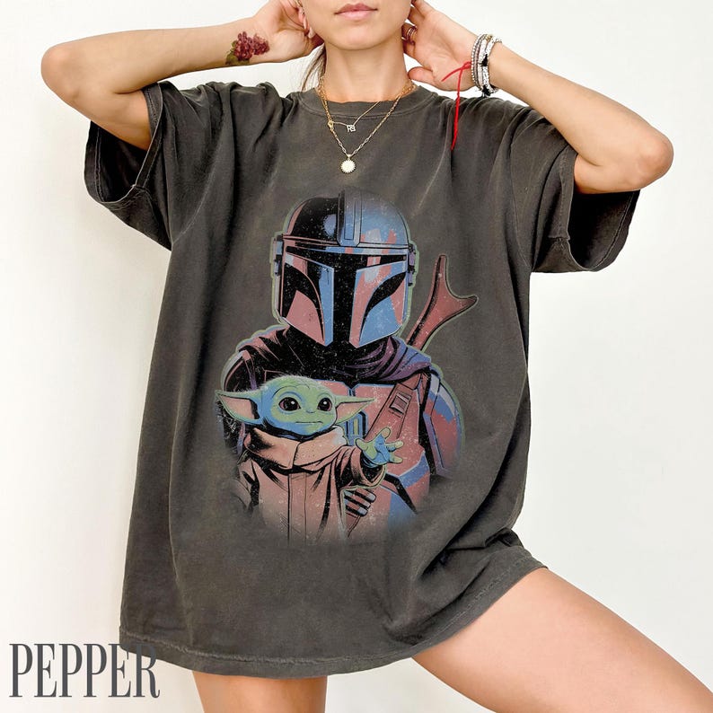 Puede incluir: Una camiseta de manga corta gris carb&oacute;n con un estampado gr&aacute;fico de un guerrero con casco en tonos azules y rojos, junto a una peque&ntilde;a criatura verde con t&uacute;nica beige. El dise&ntilde;o tiene una est&eacute;tica desgastada de estilo vintage.