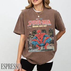 May include: Espresso-colored t-shirt featuring a Marvel Studios Spider-Man graphic. The design includes the words "Spider-Man Brand New Day" in red and white, with an illustration of Spider-Man in a city setting.
