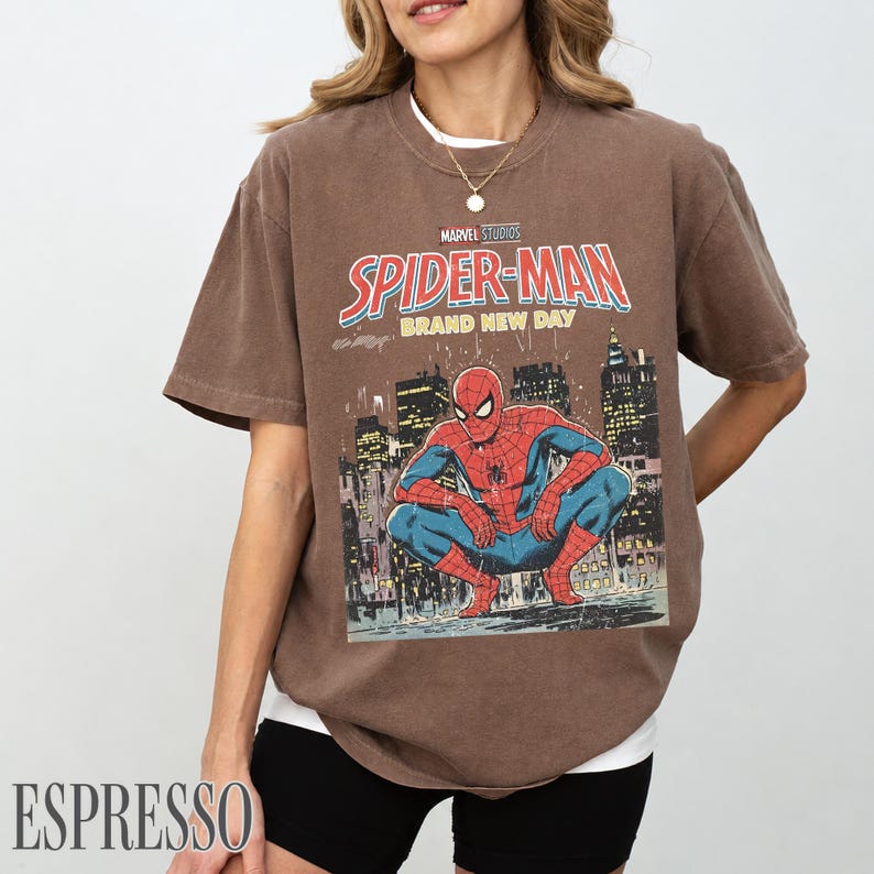 May include: Espresso-colored t-shirt featuring a Marvel Studios Spider-Man graphic. The design includes the words "Spider-Man Brand New Day" in red and white, with an illustration of Spider-Man in a city setting.