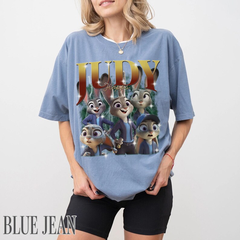 May include: A light blue t-shirt featuring a graphic of Judy Hopps from the animated film Zootopia. The design includes the name "Judy" in gold lettering and images of the character. The words "BLUE JEAN" are printed at the bottom.