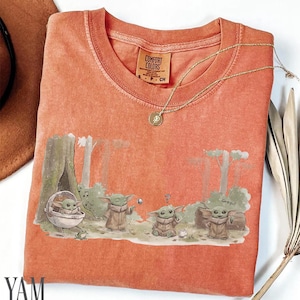 Pu&ograve; includere: T-shirt Comfort Colors color corallo con una grafica di Grogu da The Mandalorian. Il design mostra Grogu in varie pose in un ambiente forestale. Una collana d'oro con un ciondolo &egrave; appoggiata sulla maglietta. La maglietta &egrave; piegata.