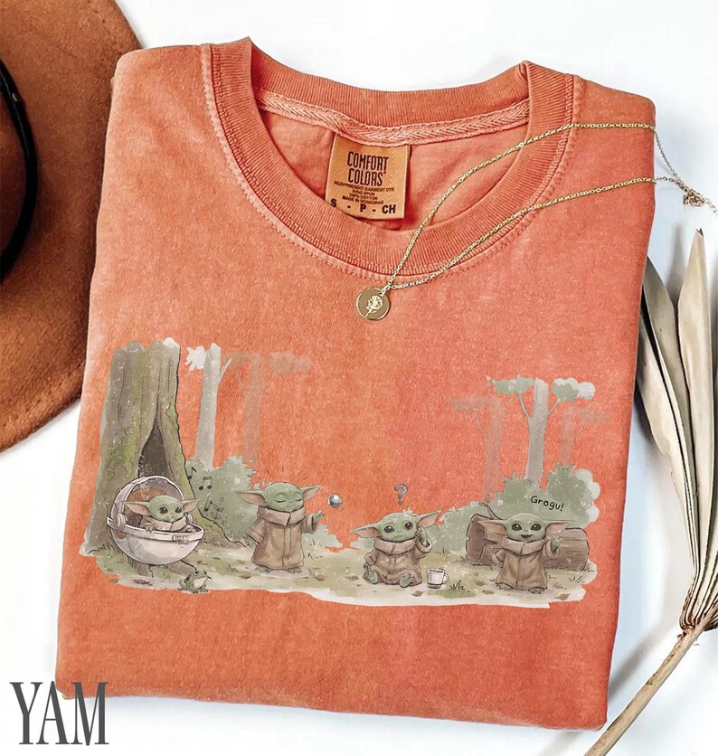 Pu&ograve; includere: T-shirt Comfort Colors color corallo con una grafica di Grogu da The Mandalorian. Il design mostra Grogu in varie pose in un ambiente forestale. Una collana d'oro con un ciondolo &egrave; appoggiata sulla maglietta. La maglietta &egrave; piegata.