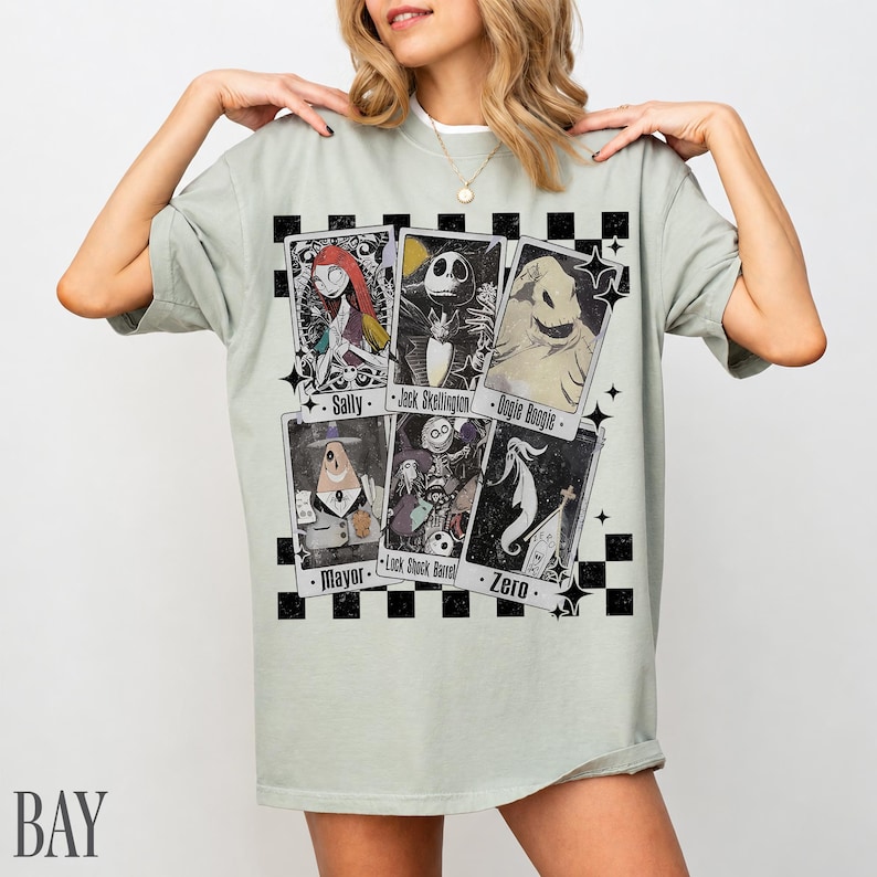 May include: A light green t-shirt featuring playing card designs with characters from The Nightmare Before Christmas. The cards include Sally, Jack Skellington, Oogie Boogie, Mayor, Lock Shock Barrel, and Zero. The shirt has a checkerboard pattern.