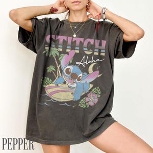 May include: Dark gray t-shirt featuring a graphic of Stitch surfing with the word "Stitch Aloha" in a retro style. The design includes palm trees, a crescent moon, and tropical flowers. The shirt has a vintage, worn look.