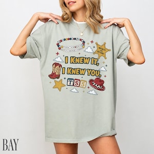 May include: A sage green t-shirt featuring a graphic print with the text I KNEW IT, I KNEW YOU, TS5, and INFINITY AND BEYOND. The design includes a beaded bracelet, a gold sheriff star, red cowboy boots, a red cowboy hat, and small clouds.