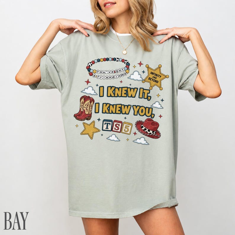 May include: A sage green t-shirt featuring a graphic print with the text I KNEW IT, I KNEW YOU, TS5, and INFINITY AND BEYOND. The design includes a beaded bracelet, a gold sheriff star, red cowboy boots, a red cowboy hat, and small clouds.