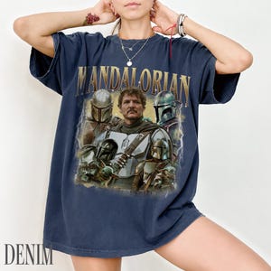 May include: Navy blue t-shirt with the gold text "MANDALORIAN" above a graphic of characters from the show. The graphic includes images of the Mandalorian, and other characters. The word "DENIM" is printed at the bottom.