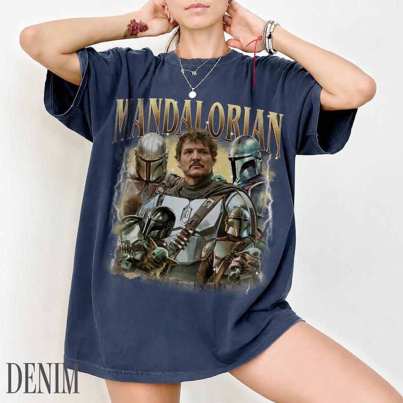 May include: Navy blue t-shirt with the gold text "MANDALORIAN" above a graphic of characters from the show. The graphic includes images of the Mandalorian, and other characters. The word "DENIM" is printed at the bottom.