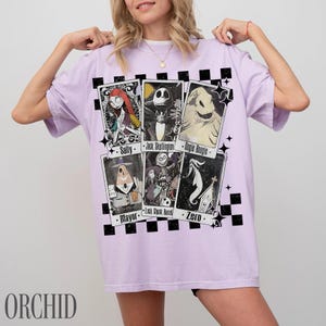 May include: A light purple t-shirt featuring character cards from The Nightmare Before Christmas. The cards include Sally, Jack Skellington, Oogie Boogie, Mayor, Lock, Shock, Barrel, and Zero. The word "ORCHID" is printed at the bottom.