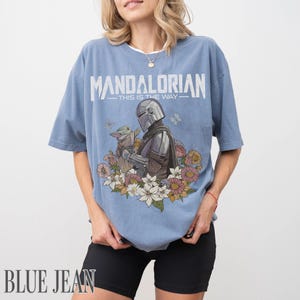 May include: A pale blue t-shirt with the word "MANDALORIAN" printed across the top. Below is an illustration of the Mandalorian and Grogu with flowers. The text "THIS IS THE WAY" is below the word.