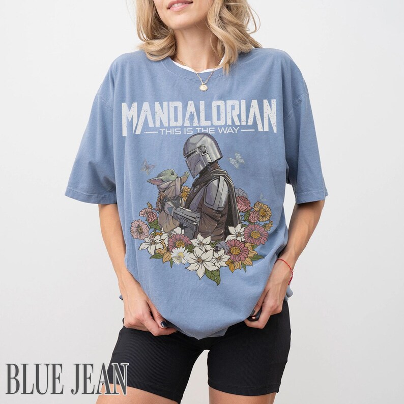 May include: A pale blue t-shirt with the word "MANDALORIAN" printed across the top. Below is an illustration of the Mandalorian and Grogu with flowers. The text "THIS IS THE WAY" is below the word.