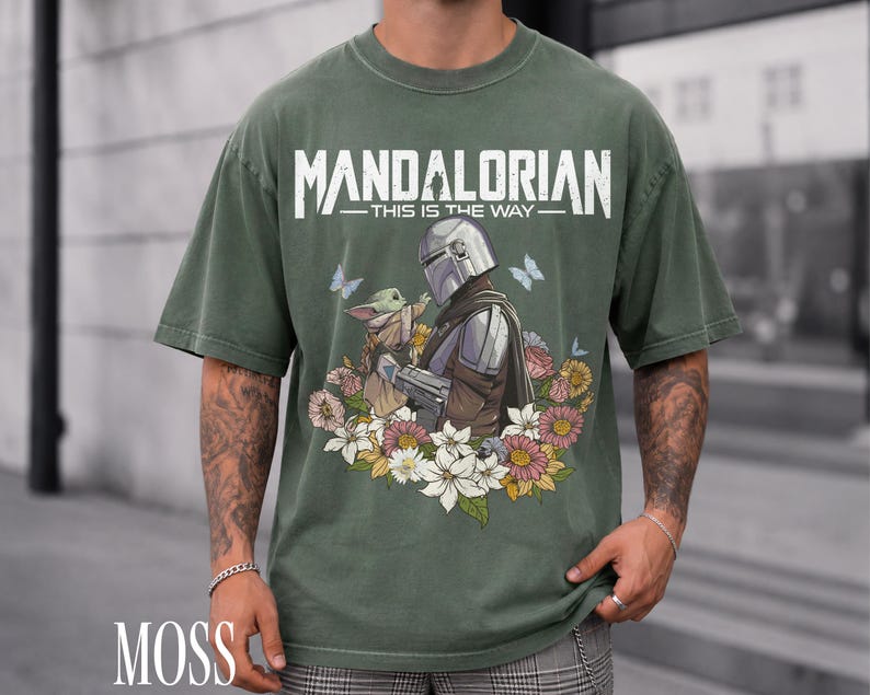 May include: Moss green t-shirt with a graphic of the Mandalorian and Grogu amidst flowers and butterflies. The text "MANDALORIAN - THIS IS THE WAY" is printed across the top. The shirt has short sleeves and a relaxed fit.