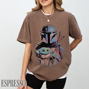 Puede incluir: Una camiseta marr&oacute;n de manga corta con un estampado gr&aacute;fico de un personaje con casco y armadura azul y gris detr&aacute;s de una peque&ntilde;a criatura verde de orejas grandes. La camiseta tiene cuello redondo y un corte relajado con dise&ntilde;o desgastado.