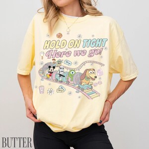 May include: Yellow t-shirt with a colorful graphic featuring Disney characters on a roller coaster, with the text "HOLD ON TIGHT Here we go!". The design includes Mickey Mouse, a Stormtrooper, and Slinky Dog.