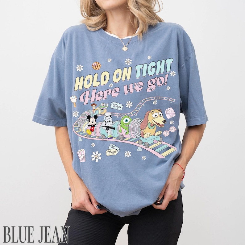 May include: A light blue t-shirt with a colorful graphic featuring Disney characters on a roller coaster, with the text "HOLD ON TIGHT Here we go!". The shirt has short sleeves and a relaxed fit.