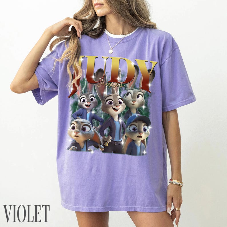 May include: Violet-colored t-shirt featuring a graphic of Judy Hopps from Zootopia. The design includes the word "JUDY" in gold and red, with images of Judy and other characters. The word "VIOLET" is printed at the bottom.