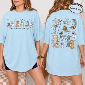 Puede incluir: Camiseta azul claro con ilustraciones de perros. La parte delantera presenta una fila de diferentes razas de perros con la frase "Life is better with dogs." La parte trasera de la camiseta muestra m&aacute;s dibujos de perros. La camiseta tiene mangas cortas y un corte holgado.