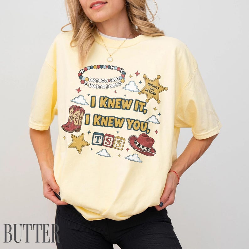 May include: A pale yellow t-shirt featuring a graphic print with the text "I KNEW IT, I KNEW YOU," "TS5," and "INFINITY AND BEYOND." The design includes illustrations of a beaded bracelet, a cowboy boot, a cowboy hat, stars, and clouds.