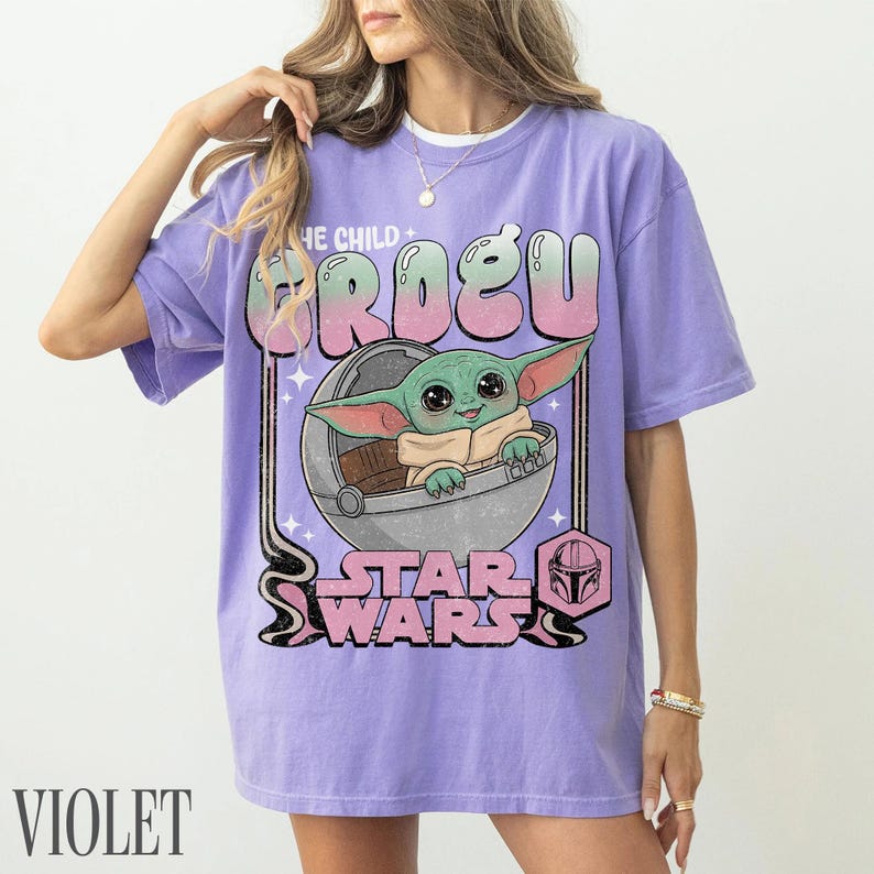 May include: A light purple short-sleeve t-shirt featuring a graphic of Grogu from Star Wars inside a floating pod. The text reads THE CHILD GROGU and STAR WARS in a retro font. The design includes pink and green gradient lettering and a small icon.