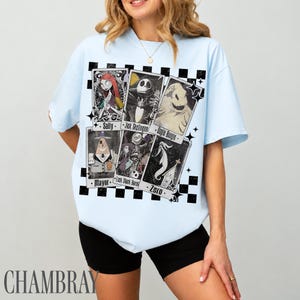 May include: Light blue t-shirt featuring a graphic of playing cards with characters from The Nightmare Before Christmas. The cards include Sally, Jack Skellington, Oogie Boogie, Mayor, Lock Shock Barrel, and Zero. The shirt has a black and white checkered pattern.