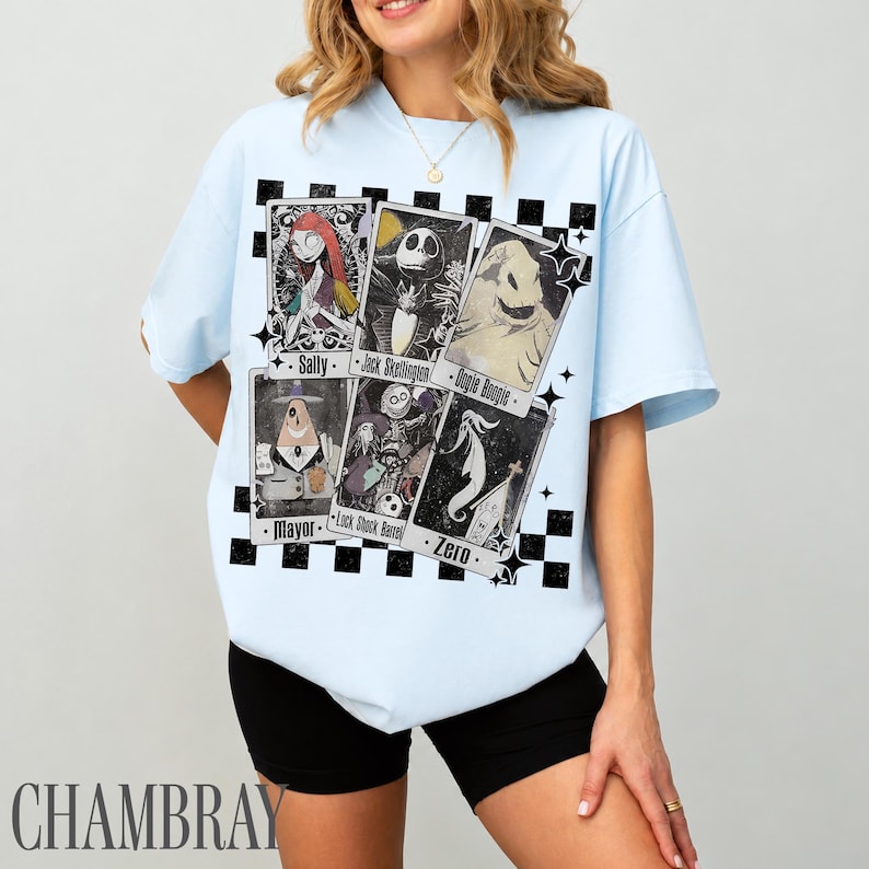 May include: Light blue t-shirt featuring a graphic of playing cards with characters from The Nightmare Before Christmas. The cards include Sally, Jack Skellington, Oogie Boogie, Mayor, Lock Shock Barrel, and Zero. The shirt has a black and white checkered pattern.