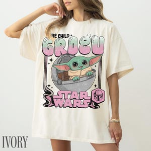 May include: An ivory t-shirt featuring a graphic print of Grogu, also known as The Child, sitting in a floating pod. The design includes the text THE CHILD GROGU and STAR WARS in a retro, distressed pink and green font, with a small helmet icon.
