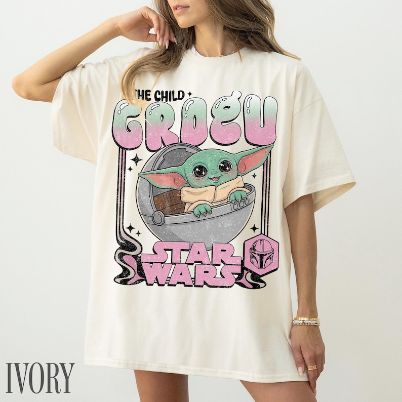May include: An ivory t-shirt featuring a graphic print of Grogu, also known as The Child, sitting in a floating pod. The design includes the text THE CHILD GROGU and STAR WARS in a retro, distressed pink and green font, with a small helmet icon.