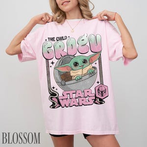 May include: A light pink short-sleeve t-shirt featuring a graphic of the character Grogu in a floating pod. The text reads THE CHILD GROGU and STAR WARS, with a small Mandalorian helmet icon. The design uses a gradient of green and pink colors.