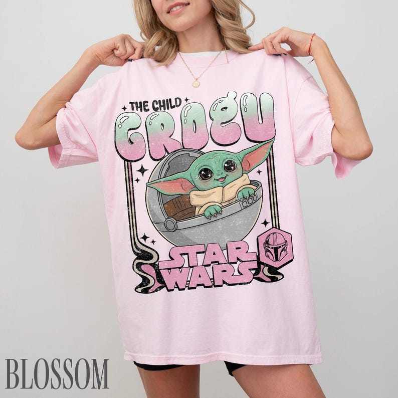 May include: A light pink short-sleeve t-shirt featuring a graphic of the character Grogu in a floating pod. The text reads THE CHILD GROGU and STAR WARS, with a small Mandalorian helmet icon. The design uses a gradient of green and pink colors.