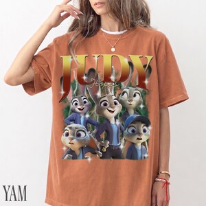 May include: Burnt orange t-shirt featuring a graphic of the character Judy Hopps from the animated film Zootopia. The design includes the word "JUDY" in gold lettering and several cartoon bunnies. The shirt has short sleeves and a crew neck.