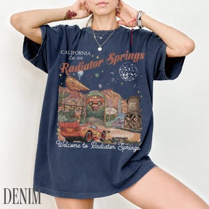 May include: A dark blue short-sleeve t-shirt featuring a graphic print of the Radiator Springs landscape from the movie Cars. The design includes a red race car, desert rock formations, a water tower, and the text California Est. 2012 Radiator Springs.