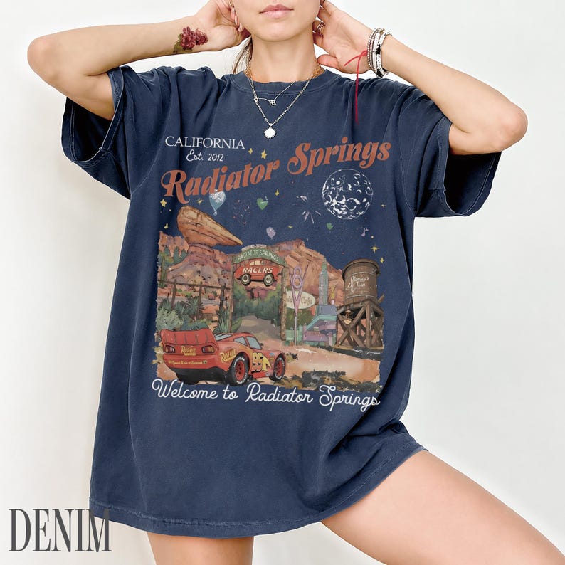 May include: A dark blue short-sleeve t-shirt featuring a graphic print of the Radiator Springs landscape from the movie Cars. The design includes a red race car, desert rock formations, a water tower, and the text California Est. 2012 Radiator Springs.