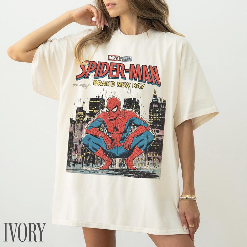 May include: Ivory-colored t-shirt featuring a comic book-style illustration of Spider-Man. The graphic includes the words "Marvel Studios Spider-Man Brand New Day" above the superhero. The shirt has short sleeves and a relaxed fit.