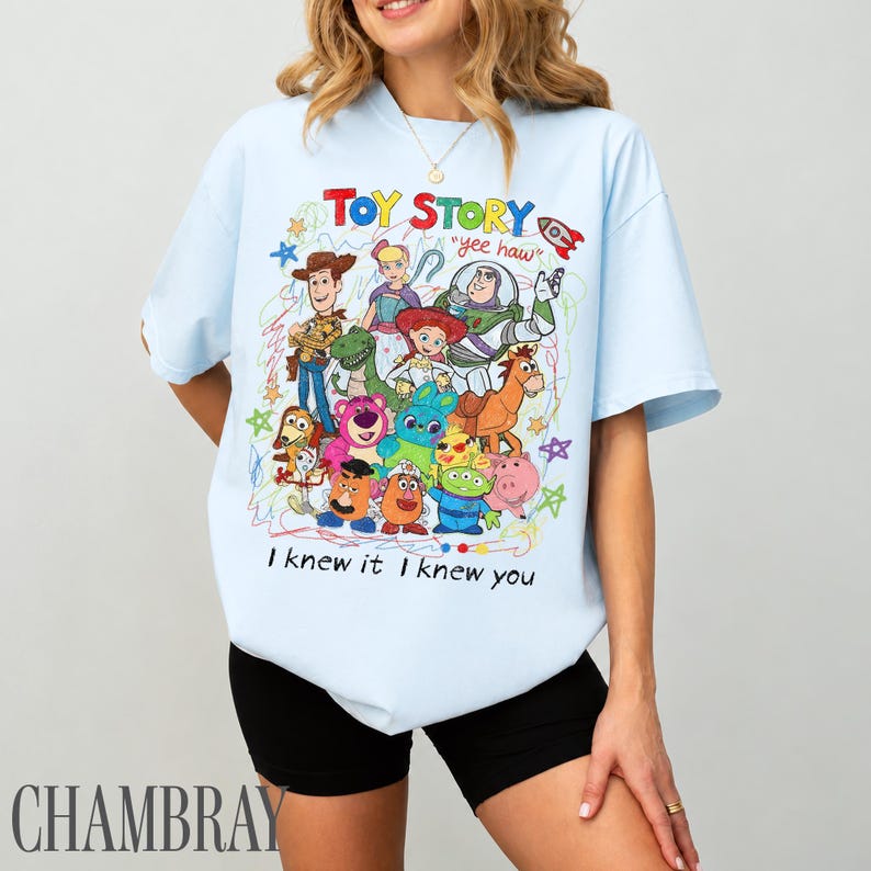 May include: A light blue t-shirt featuring a colourful graphic print of Toy Story characters including Woody, Buzz Lightyear, Jessie, and others. The design includes the text "TOY STORY", "yee haw", and "I knew it I knew you" surrounded by hand-drawn style stars.