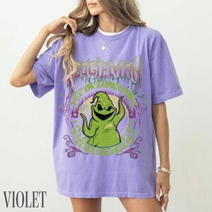 May include: Violet t-shirt with a green Oogie Boogie graphic and the text "OOGIE BOOGIE" above and "WHAT HAVE WE HERE" below. The shirt has a crew neck and short sleeves.