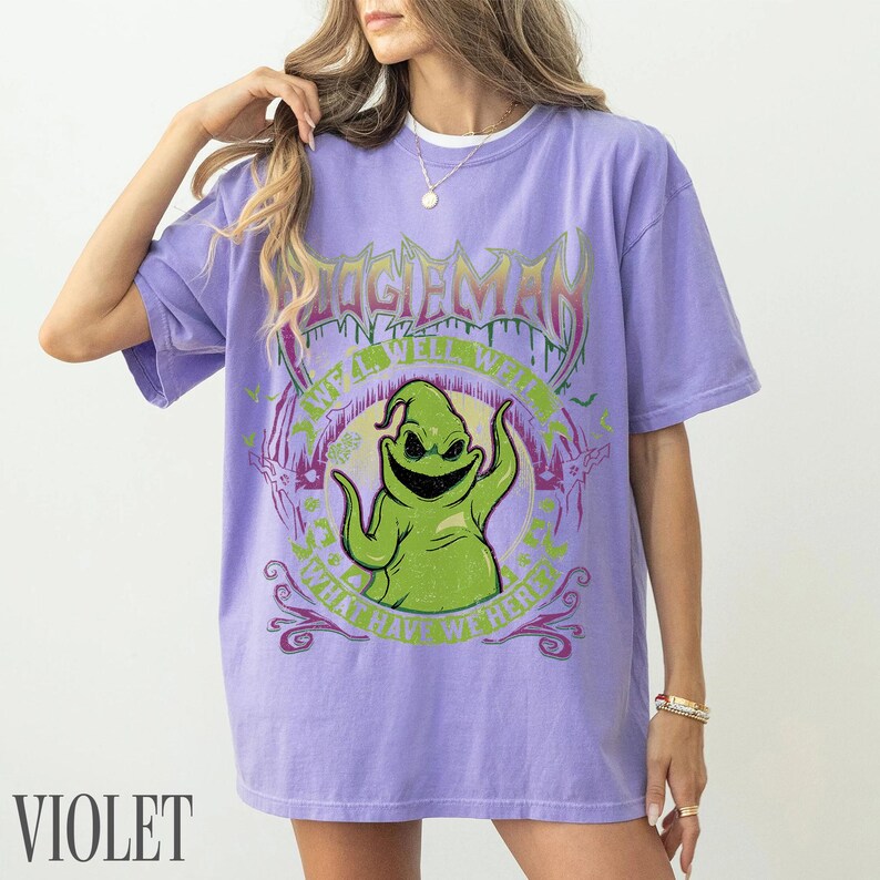 May include: Violet t-shirt with a green Oogie Boogie graphic and the text "OOGIE BOOGIE" above and "WHAT HAVE WE HERE" below. The shirt has a crew neck and short sleeves.