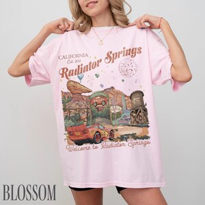 May include: A light pink t-shirt featuring a graphic print of Radiator Springs with a red race car, desert rock formations, and a water tower. The text reads California Est. 2012, Radiator Springs, and Welcome to Radiator Springs in brown script.