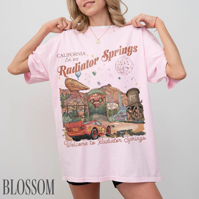 May include: A light pink t-shirt featuring a graphic print of Radiator Springs with a red race car, desert rock formations, and a water tower. The text reads California Est. 2012, Radiator Springs, and Welcome to Radiator Springs in brown script.