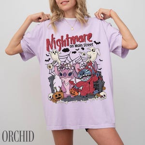 May include: A light purple t-shirt featuring a graphic print with the text Nightmare on Main Street. The design includes two cartoon characters, one pink and one blue, surrounded by spiderwebs, ghosts, bats, pumpkins, and skulls.