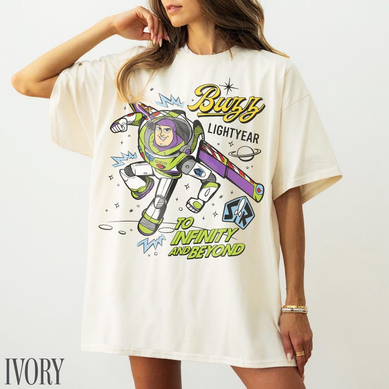 Pode incluir: Camiseta de cor marfim com um desenho de Buzz Lightyear em uma pose din&acirc;mica. O design inclui as palavras "Buzz Lightyear" e "To Infinity and Beyond". A camiseta tem mangas curtas e gola redonda.