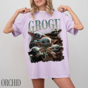 K&ouml;nnte beinhalten: Ein helllila T-Shirt mit einer Grafik von Grogu aus The Mandalorian. Das Design enth&auml;lt mehrere Bilder von Grogu mit dem Wort "GROGU" dar&uuml;ber. Das Wort "ORCHID" ist unten auf dem Shirt aufgedruckt.